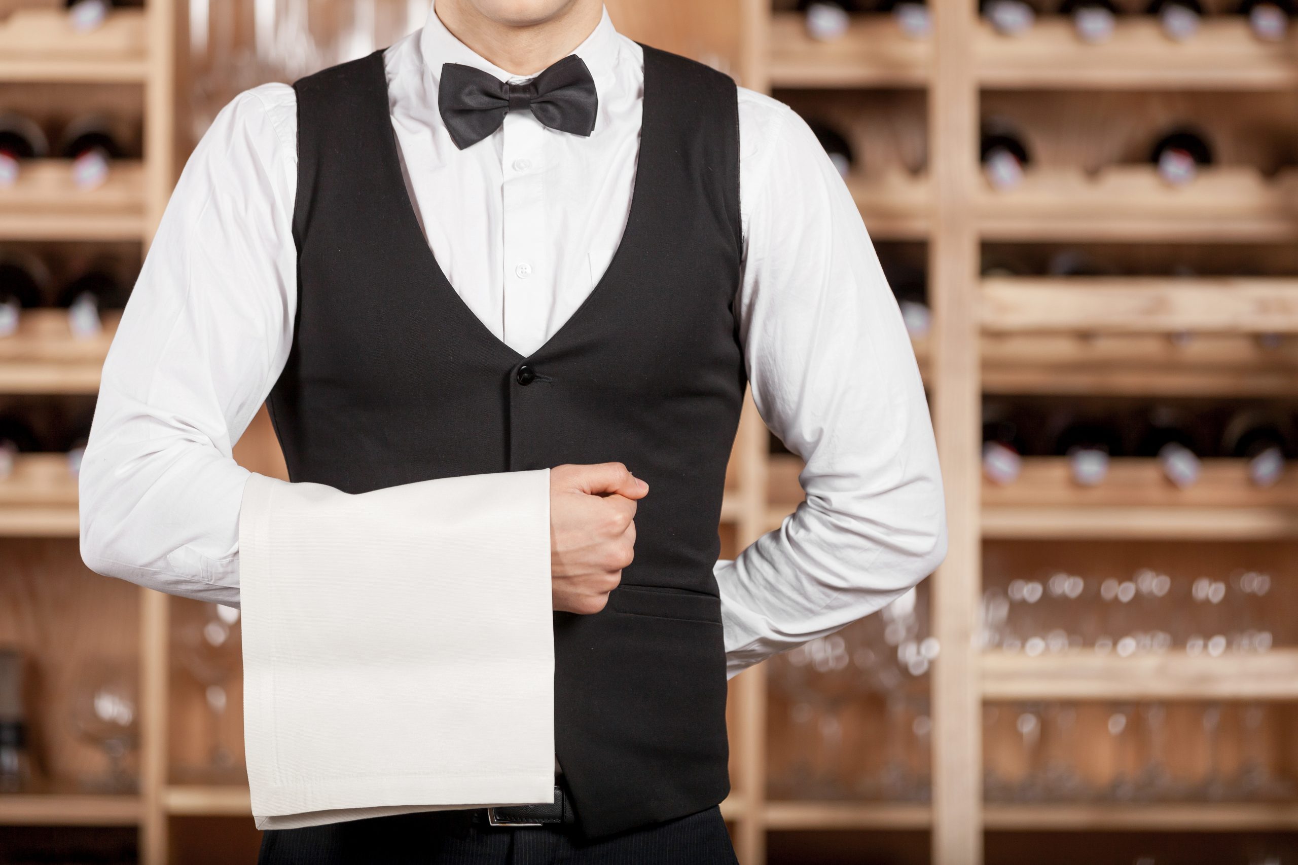 Confident waiter. Cropped image of confident young waiter standing in front of wine shelf and holding a towel on his hand